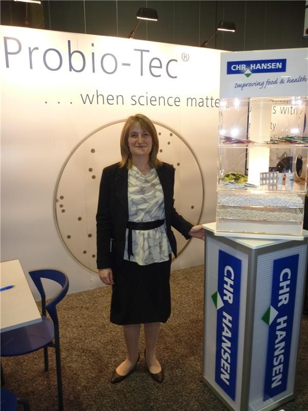 A woman in a dark jacket and patterned blouse stands beside a CHR Hansen display pedestal in front of a Probio-Tec backdrop reading "when science matters." A table and blue chair are at left.