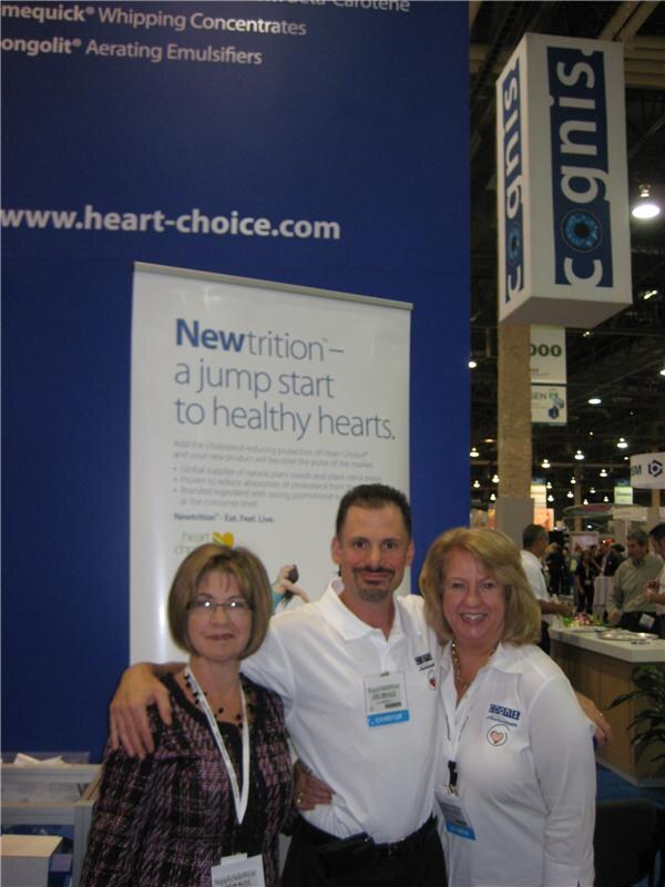 Two women and a man with arms around each other pose in front of a Cognis booth with a Newtrition Heart Choice banner reading "a jump start to healthy hearts." A hanging Cognis sign is above.