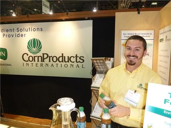 A smiling bearded man in a yellow CornProducts shirt holds a small bottle at a table with more bottles and a pitcher, in front of a Corn Products International sign.
