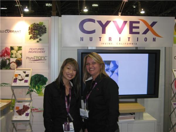 Two women in black tops with lanyards stand in front of a Cyvex Nutrition, Irvine, California sign and a screen. A panel at left features PhytoTropic, and brochure racks stand nearby.