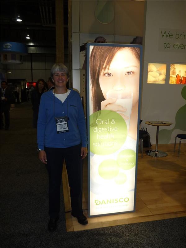 A gray-haired woman in a blue cardigan with an exhibitor badge stands beside a lit Danisco panel showing a woman drinking milk and the text "Oral & digestive health solutions."