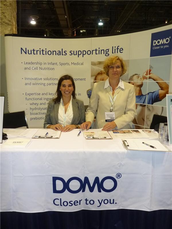 Two women in blazers sit smiling behind a table with a white DOMO cloth reading 'Closer to you.' A banner behind them reads 'Nutritionals supporting life' with the DOMO logo.