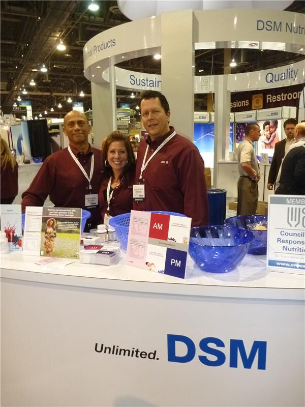 Two men and a woman in maroon shirts and lanyards stand behind a white DSM counter with brochures and blue bowls on top. The counter reads 'Unlimited. DSM.'