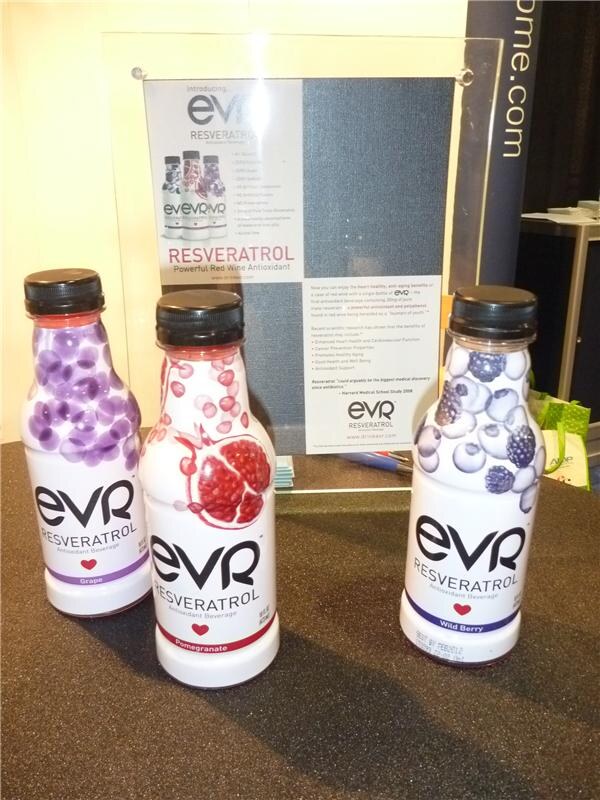 Three white EVR Resveratrol antioxidant beverage bottles with grape, pomegranate and wild berry artwork stand on a dark table in front of an EVR Resveratrol information sign.