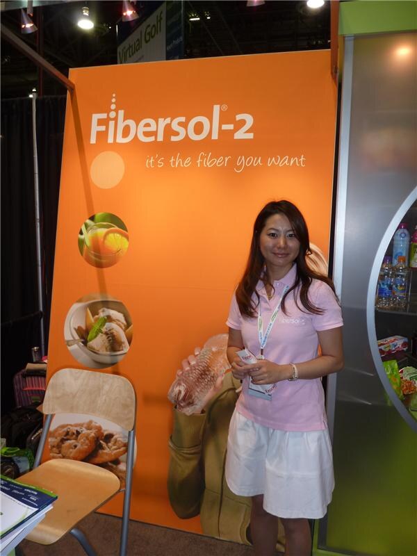 A woman in a pink polo and white skirt stands holding a package in front of an orange Fibersol-2 banner reading 'it's the fiber you want'.