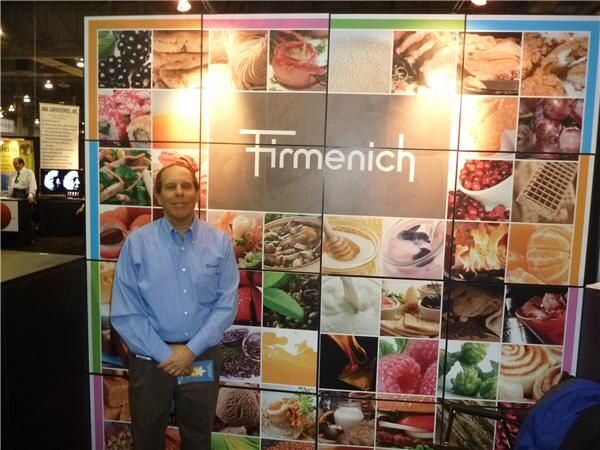 A man in a blue shirt stands in front of a large Firmenich video wall made up of food images including berries, vegetables, grains and desserts.