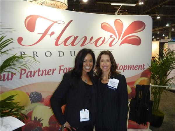 Two smiling women in dark clothing stand in front of a Flavor Producers banner with fruit images. One wears an exhibitor badge.