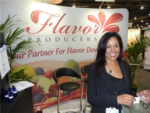 A smiling woman with long dark hair in a black jacket stands at a counter in front of a Flavor Producers banner with fruit images and bar stools.