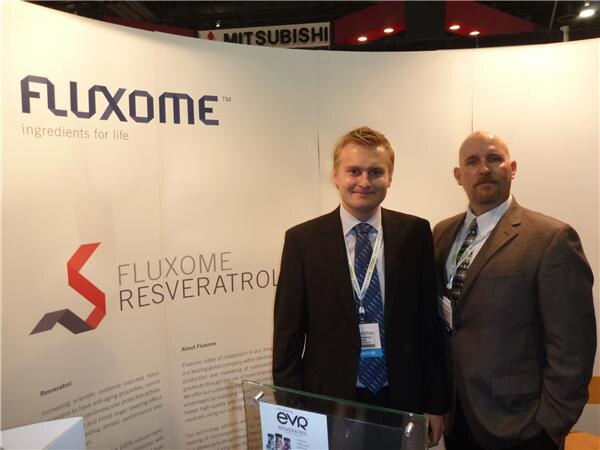 Two men in suits and lanyards stand in front of a Fluxome Resveratrol banner. A Mitsubishi sign hangs above and an EVR Resveratrol display sits in the foreground.