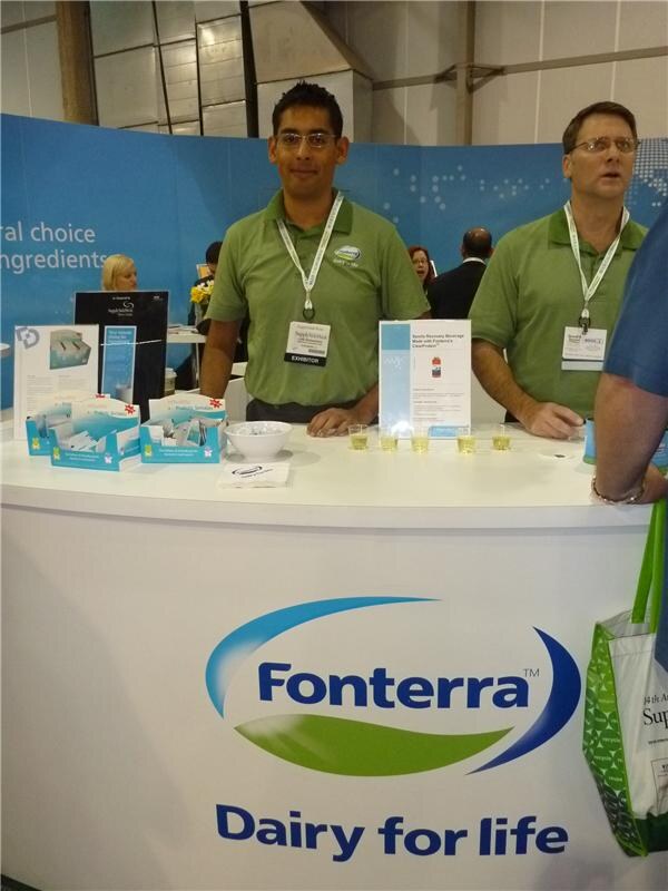 Two men in green Fonterra polo shirts and exhibitor badges stand behind a curved white booth counter with the Fonterra Dairy for life logo, small sample cups, brochures and a bowl on top.