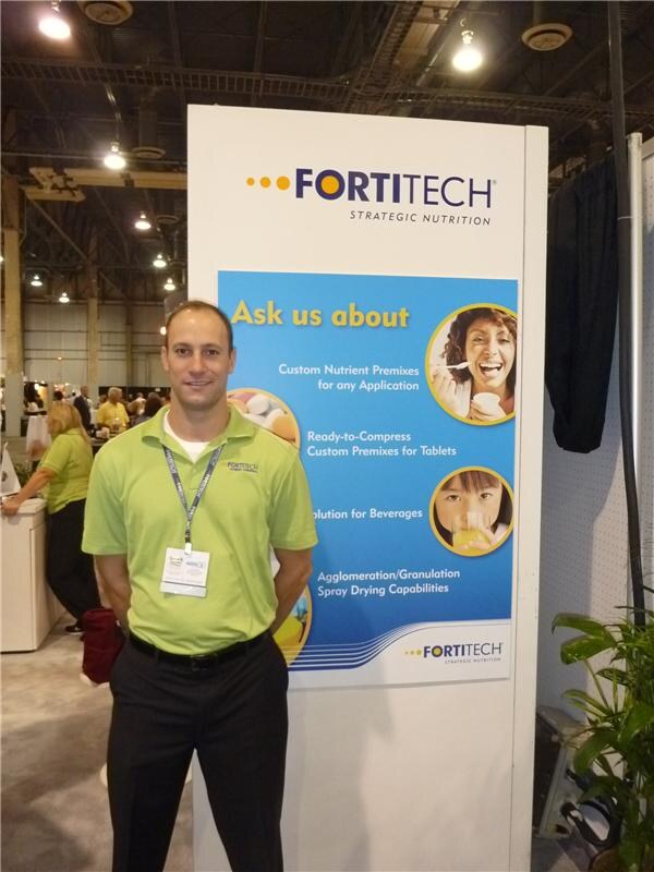 A smiling man in a green Fortitech polo and lanyard stands in front of a Fortitech Strategic Nutrition sign reading Ask us about custom nutrient premixes, with a plant beside it.