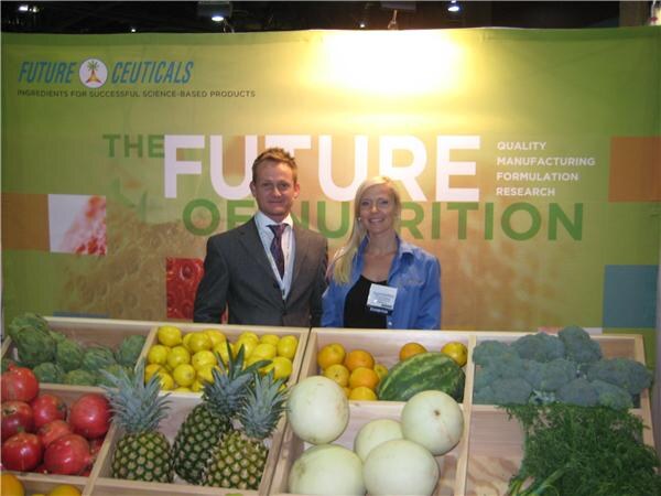 A man in a gray suit and a woman in a blue jacket stand behind crates of pineapples, tomatoes, lemons, oranges and melons in front of a Future Ceuticals banner reading The Future of Nutrition.