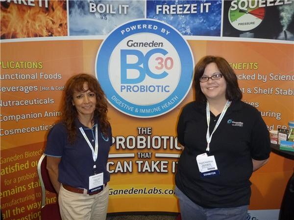 Two smiling women with exhibitor badges stand in front of an orange Ganeden banner with a round Powered by Ganeden BC30 Probiotic logo and the text The probiotic that can take it.