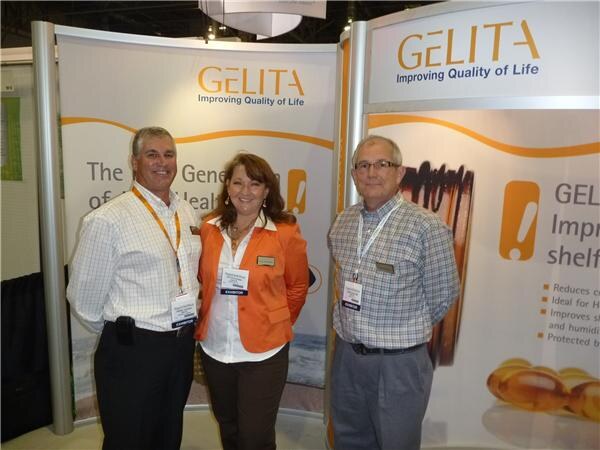 A woman in an orange jacket stands between two men in checked shirts, all wearing exhibitor badges, in front of Gelita Improving Quality of Life banners showing softgel capsules.