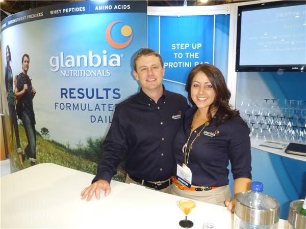 A man and a woman in dark Glanbia Nutritionals shirts lean on a white counter in front of a Glanbia banner and a Protini bar sign, with a cocktail glass and a water pitcher nearby.
