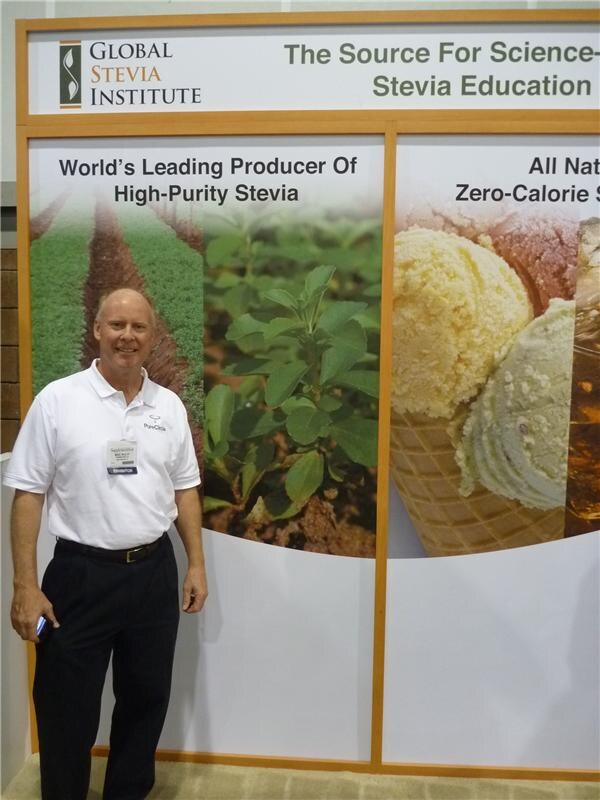 A smiling bald man in a white PureCircle polo and badge stands beside Global Stevia Institute panels showing stevia plants and ice cream, with text on high-purity stevia.