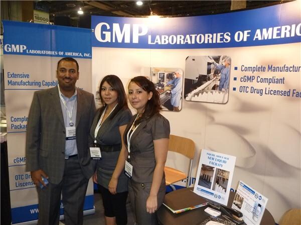 A man in a gray blazer and two women in gray outfits, all with badges, stand in a GMP Laboratories of America booth with banners about manufacturing and a brochure on the table.