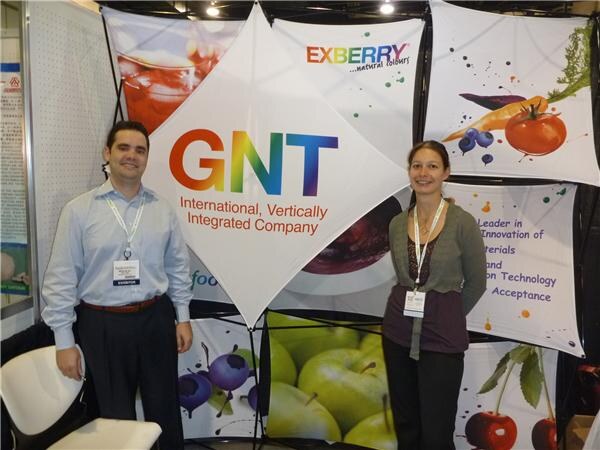 A man in a light blue shirt and a woman in a gray cardigan wear badges and stand in front of GNT and Exberry natural colours banners with images of apples, cherries and vegetables.