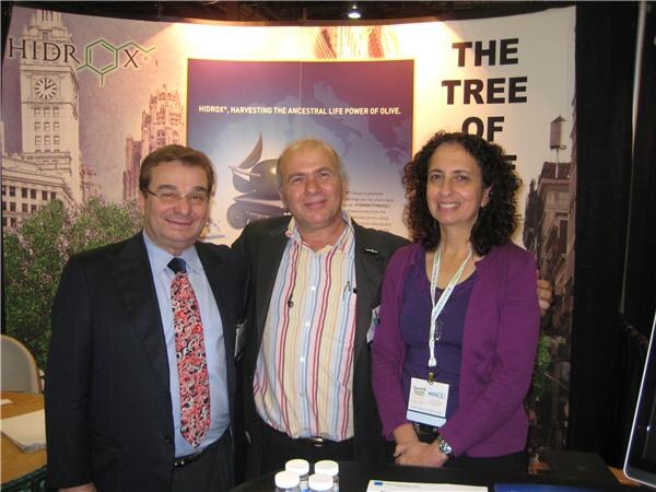 Two men and a woman stand together in a booth with Hidrox signage and a city scene backdrop. The woman wears a lanyard badge, and small white containers sit on the table in front.