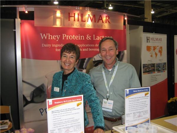 A smiling woman in a teal jacket and a man in a gray-green shirt stand behind a table with sample cups and signs in front of Hilmar Ingredients banners about whey protein and lactose.