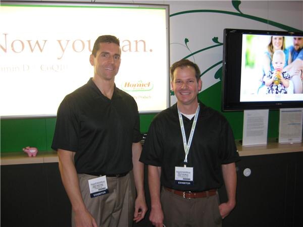 Two men in black polo shirts and exhibitor badges stand in front of a screen showing the Hormel logo and the words "Now you can." A television beside them shows a woman and a baby.