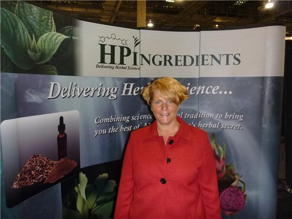 A woman with short reddish-blond hair wears a red jacket and stands in front of an HP Ingredients display reading "Delivering Herbal Science," with a dropper bottle and dried herbs pictured.