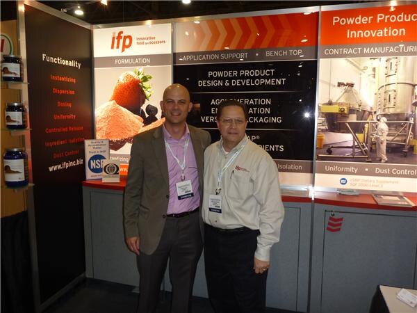 Two men stand in front of an ifp innovative food processors booth with panels on powder product innovation and an NSF logo displayed. One wears a gray blazer, the other a cream shirt.