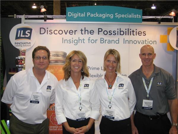 Two men and two women in ILS-branded shirts stand in front of an ILS Innovative Labeling Solutions booth with the text "Digital Packaging Specialists" and "Discover the Possibilities."
