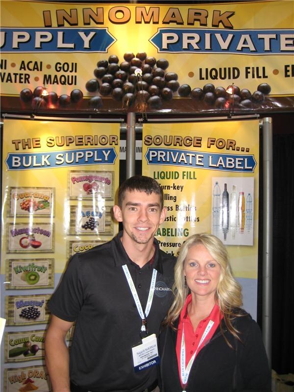 A smiling man in a black polo and a smiling woman with long blond hair in a red top stand in front of Innomark banners about bulk supply and private label sourcing.