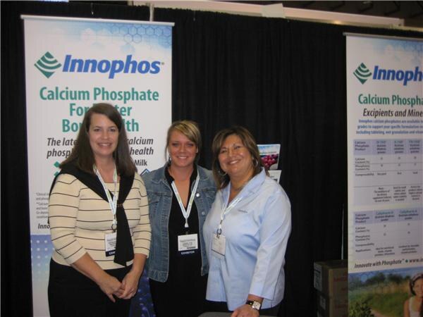 Three women wearing exhibitor badges stand together in front of Innophos banners about calcium phosphate.