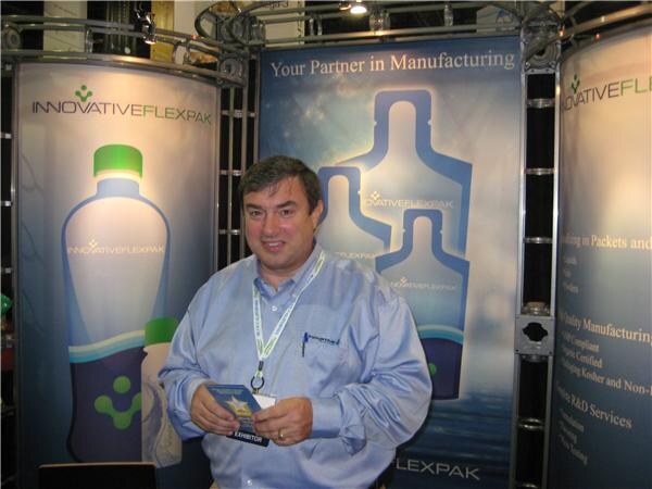 A man in a light blue button-up shirt holds a small card in front of Innovative FlexPak banners showing bottle-shaped packets and the line "Your Partner in Manufacturing."