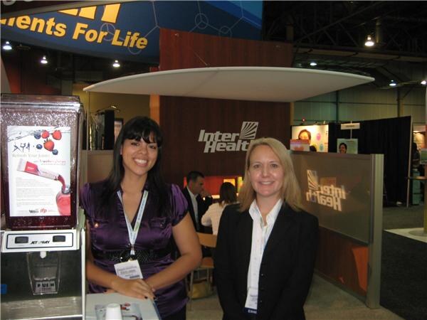 A woman in a purple top and a woman in a black blazer stand in front of InterHealth signage. A beverage dispenser with a poster is at left, and other attendees are seated behind them.
