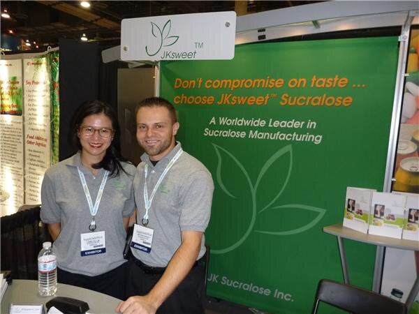 A woman with glasses and a man, both in gray polo shirts, stand in front of a green JKsweet banner reading "Don't compromise on taste... choose JKsweet Sucralose."