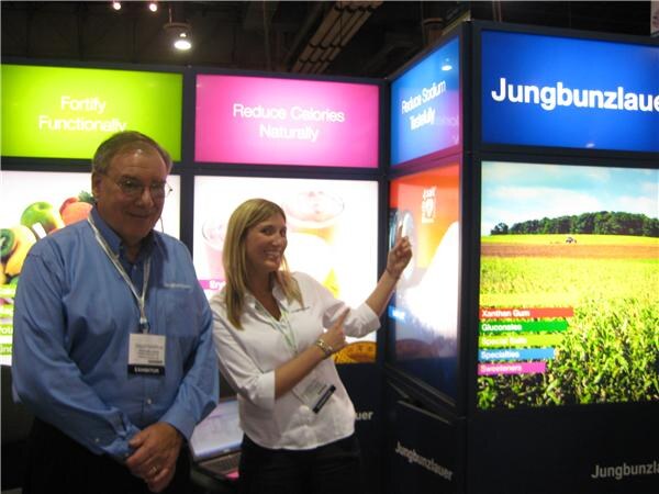 An older man in a blue shirt stands beside a woman in a white blouse who points at a lit display panel at the Jungbunzlauer booth, with panels reading "Fortify Functionally" and "Reduce Calories Naturally."