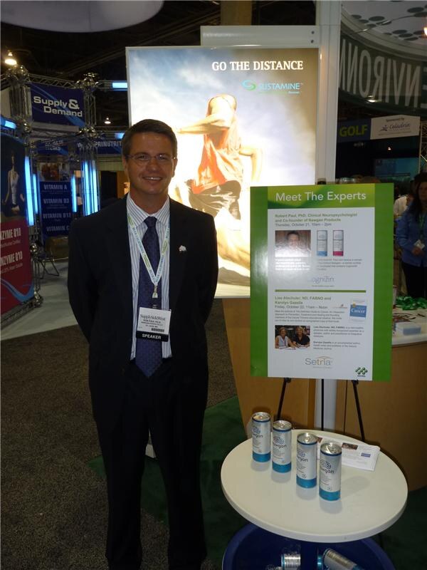 A smiling man in a dark suit and speaker badge stands beside a Sustamine "Go the Distance" display and a Meet The Experts sign for Cognizin and Setria, with Newgon cans on a table.
