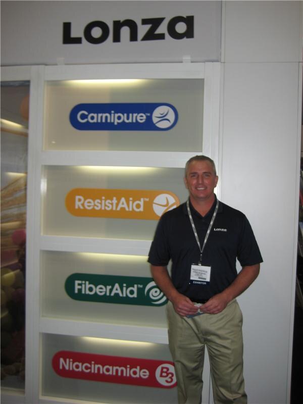 A smiling man in a dark Lonza polo and exhibitor badge stands in front of a white wall with Lonza signage and logos for Carnipure, ResistAid, FiberAid and Niacinamide.