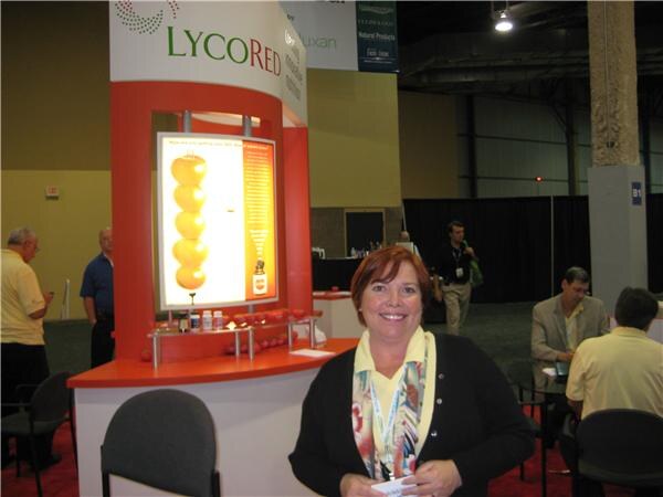 A smiling woman with short red hair and a lanyard stands in front of the LycoRed booth, which has an illuminated panel of tomatoes. Other attendees sit and stand in the background.