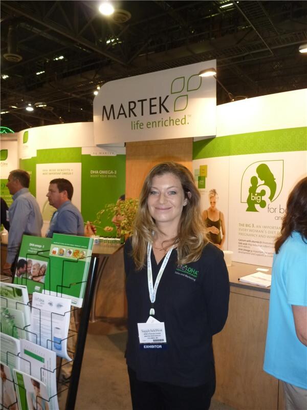 A smiling woman with long hair wears a black life's DHA shirt and exhibitor badge in front of the Martek "life enriched" booth, with brochure racks beside her and DHA omega-3 signage behind.