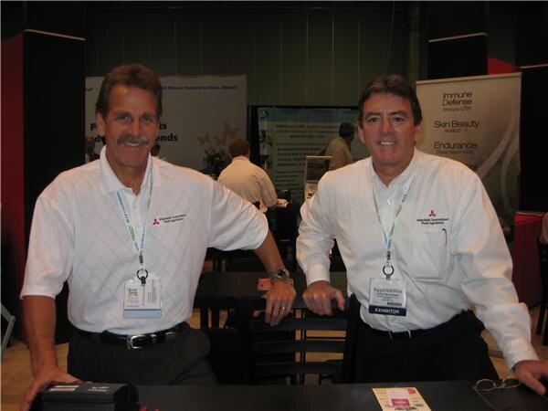 Two smiling men in white shirts with Mitsubishi International Food Ingredients logos and exhibitor badges lean on a table at the booth, with banners behind them.