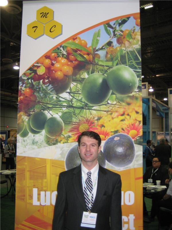 A man in a dark pinstripe suit and tie stands in front of a tall MTC banner showing green fruit, orange berries and flowers. Other attendees are seated in the background.