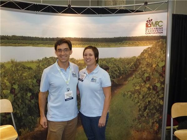 A man with glasses and a woman, both in light blue MPC polo shirts, stand in front of a Muscadine Products Corporation backdrop showing a vineyard beside a lake.