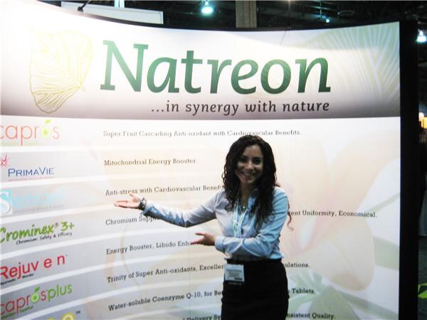 A smiling woman with long dark curly hair gestures toward a Natreon backdrop listing products including Capros, PrimaVie, Crominex 3+, Rejuven and Caprosplus.