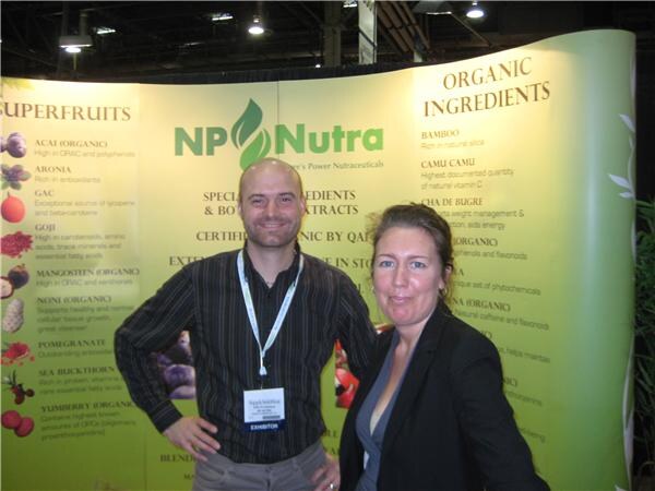 A bald man in a striped shirt with an exhibitor badge and a woman in a dark blazer stand in front of an NP Nutra backdrop listing superfruits and organic ingredients.