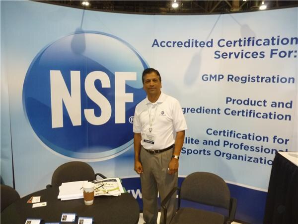 A man in a white polo shirt stands in front of a large NSF backdrop advertising accredited certification services, with a table, chairs and a coffee cup beside him.