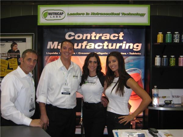 Two men in white shirts and two women in white Nutricap Labs tops pose together in front of a Contract Manufacturing banner at the Nutricap Labs booth.