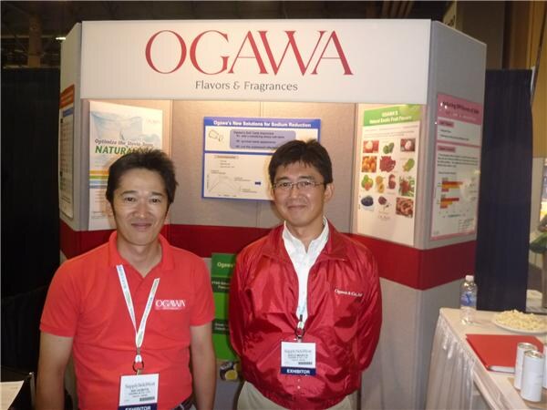 Two men in red, one in a polo and one in a jacket, wear exhibitor badges in front of the Ogawa Flavors & Fragrances booth display.