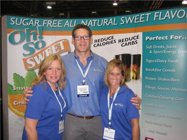 Two women and a man in blue polo shirts and exhibitor badges pose together in front of an Oh! So Sweet sugar free all natural sweet flavors banner.