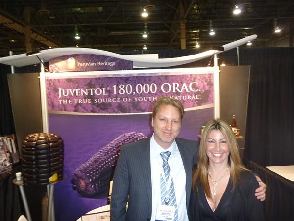 A man in a pinstripe suit and a smiling woman with long hair pose in front of a purple Juventol 180,000 ORAC banner showing purple corn at the Peruvian Heritage booth.