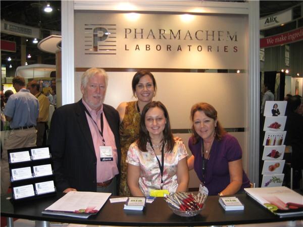 A white-haired man and three women gather behind a counter at the Pharmachem Laboratories booth, with brochures and a bowl of pens on the table.