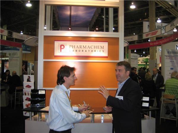 Two men talk at the Pharmachem Laboratories booth, one gesturing with his hand, in front of an orange wall with the company logo on the show floor.